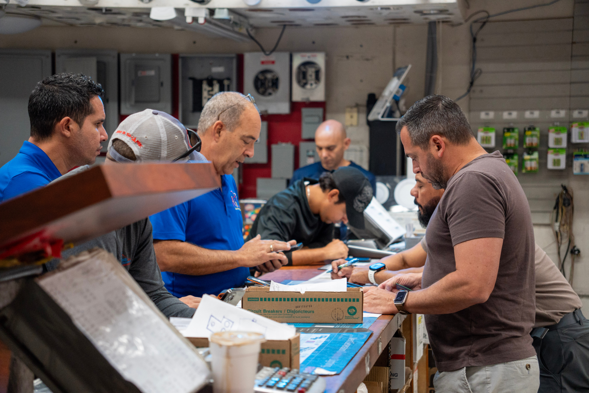 Group of men gathered around a counter in an electrical supply store, reviewing paperwork and writing notes. Boxes, tools, and electrical equipment are visible on the counter and shelves in the background.