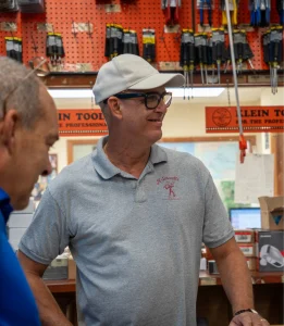 Man wearing a light gray polo shirt with a ‘Kilowatts’ logo, white cap, and glasses, smiling while standing at a counter in a hardware or electrical supply store. Tools and equipment are displayed on a red pegboard in the background.